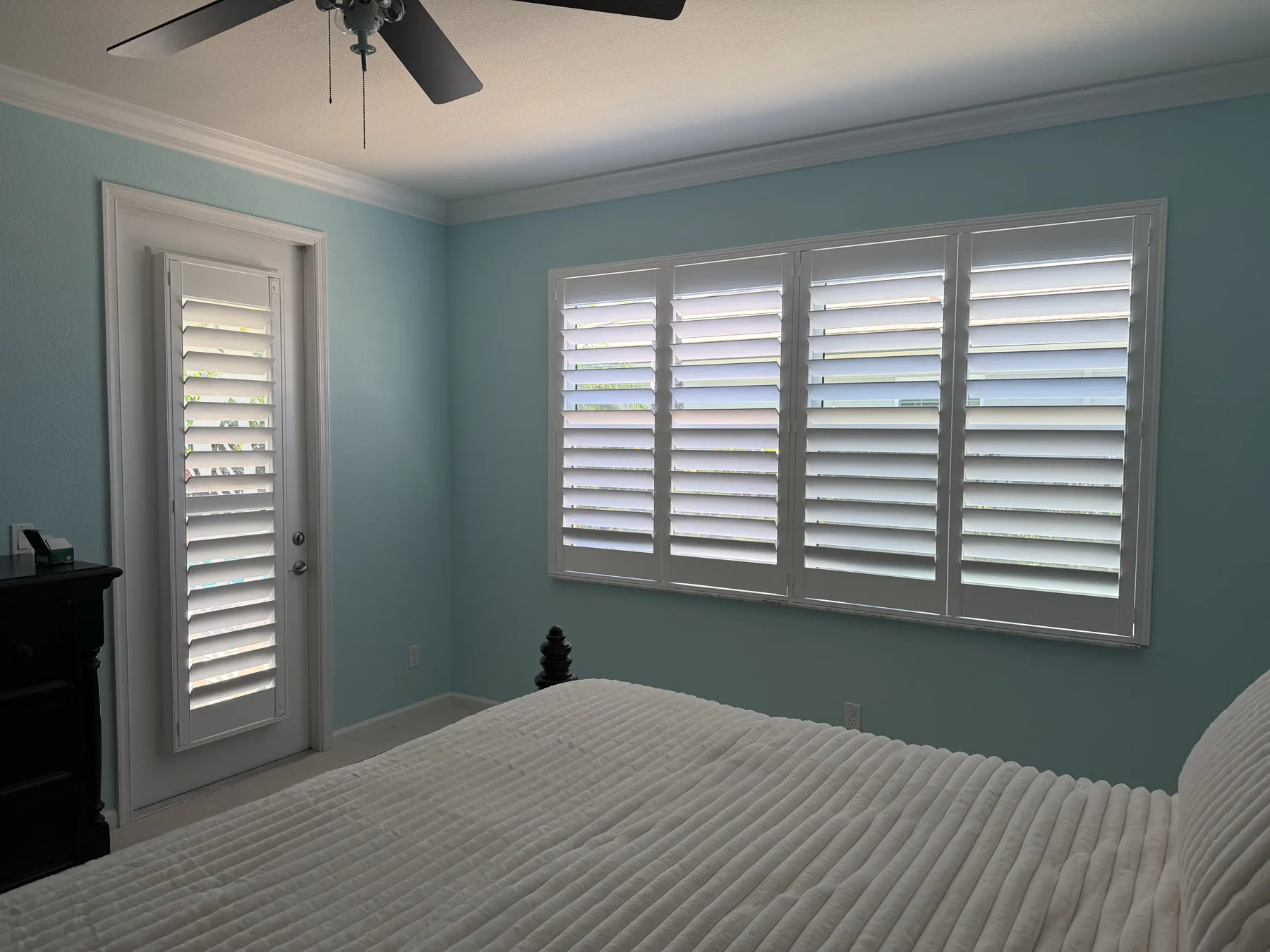 White plantation shutters across a wide window and glass door in a blue bedroom
