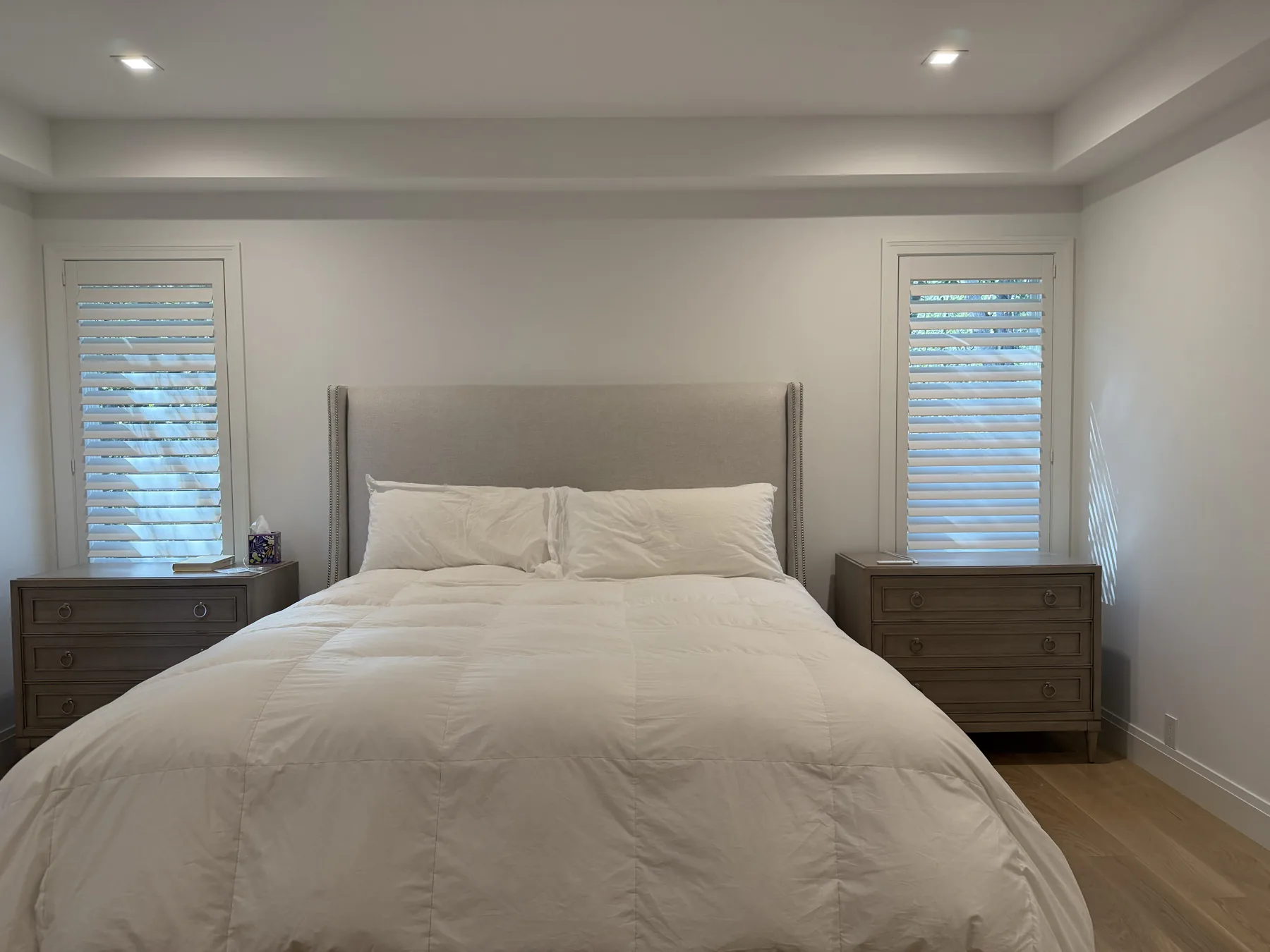 White plantation shutters on two bedroom windows beside an upholstered headboard